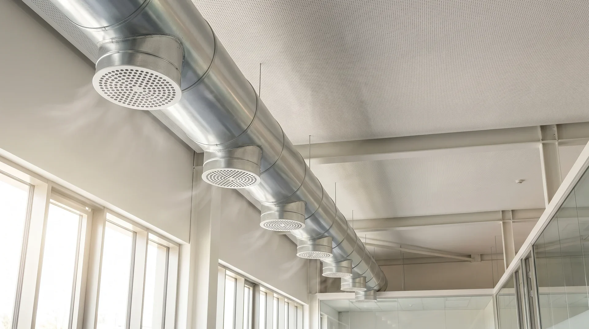 Silvrigt ventilationssystem med runda diffusorer monterat i kontorstak.